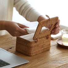 Laden Sie das Bild in den Galerie-Viewer, Person refilling a handcrafted rattan tissue box cover on a rustic wooden table, showcasing its easy-to-use hinged lid design. This eco-friendly, artisanal storage solution is perfect for enhancing natural aesthetics in home decor. Accompanied by a glass of water and a laptop, it blends functionality with style for living or working spaces.