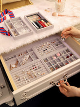 Load image into Gallery viewer, Elegant jewelry storage solution featuring stackable gray velvet trays inside a luxurious drawer, showcasing a variety of accessories like earrings, rings, bracelets, and pendants. The setup also includes a neatly organized vanity with makeup palettes, lipsticks, and a soft faux fur overlay. A hand is seen holding a teardrop earring, emphasizing the accessibility and meticulous design of the organizer, perfect for maintaining a clutter-free and stylish dressing area.